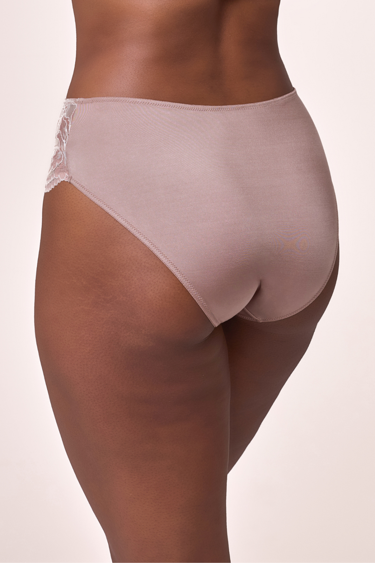 Rear view of a model wearing high-waisted, semi-sheer underwear in a dusty rose color with a lace side detail, set against a solid light beige background. Shush Lingerie